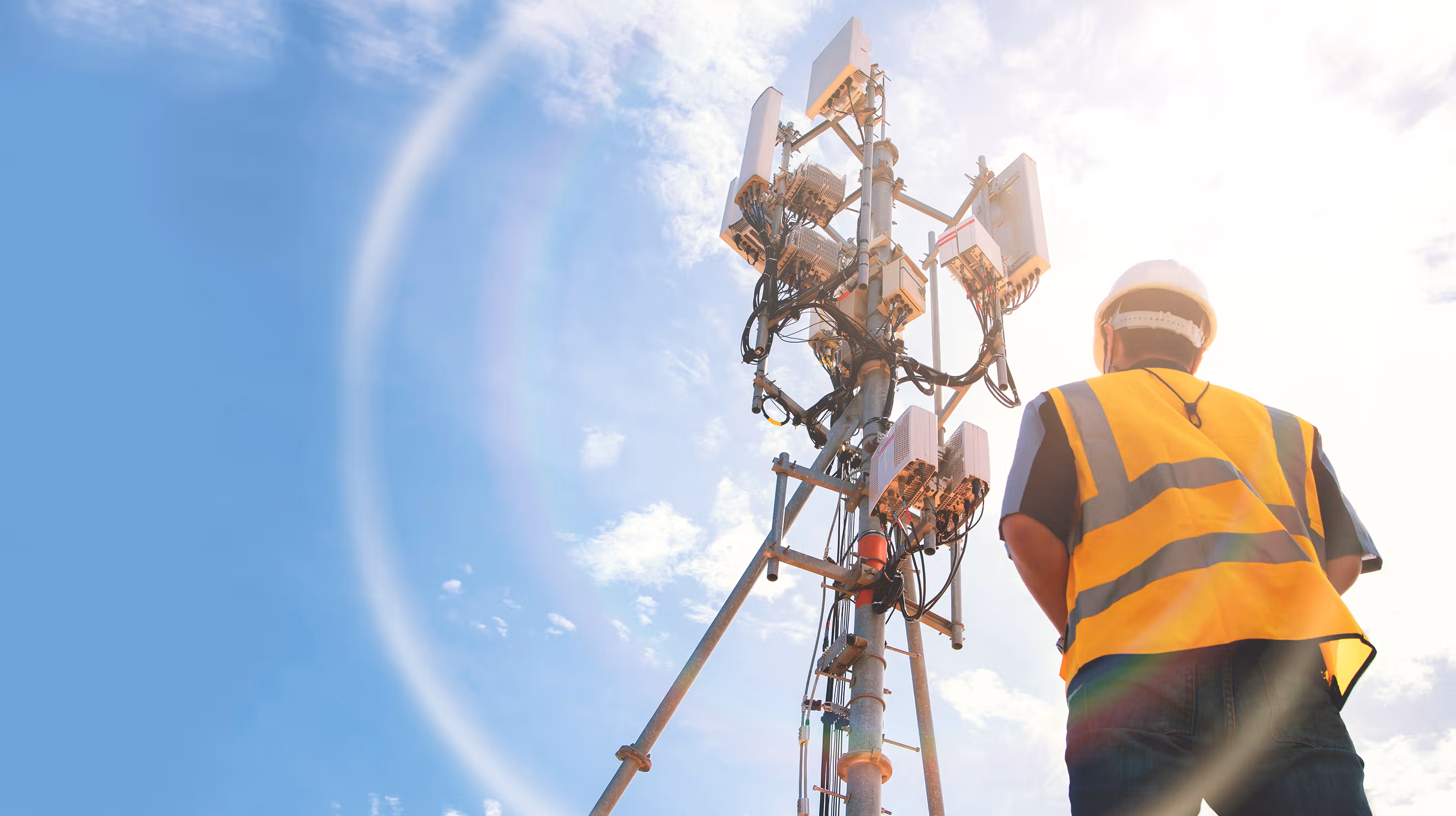 A worker wearing a safety vest staring up at a 5G tower.